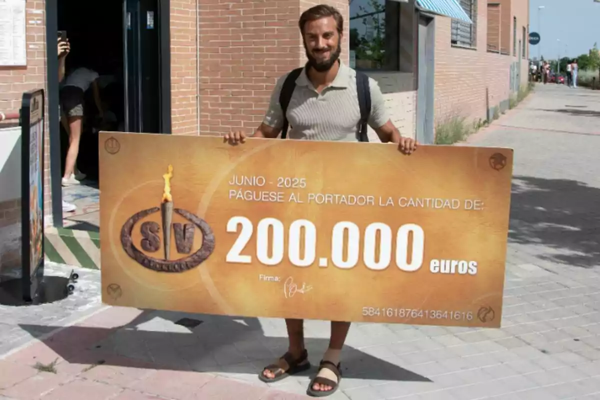 Borja González somrient sosté un xec gegant de 200.000 euros al carrer davant d’un edifici de maó.