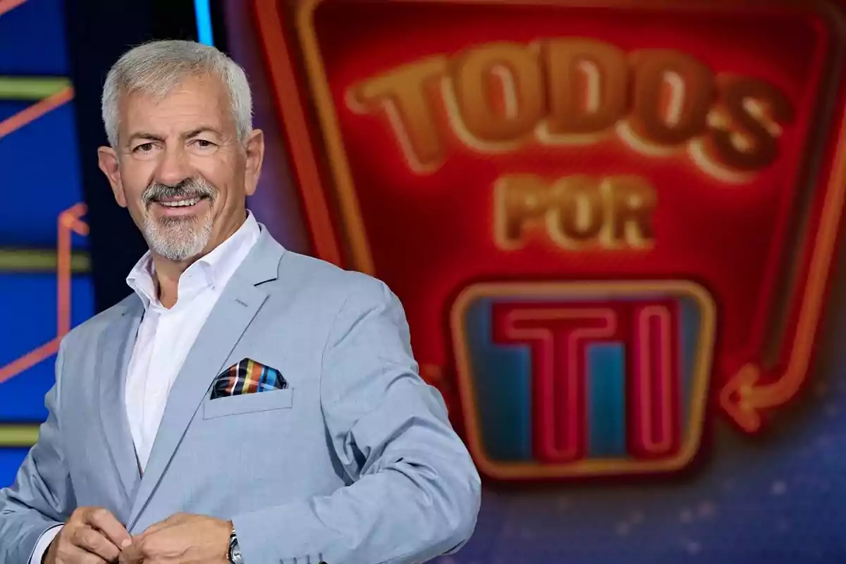 Carlos Sobera, de cabell canós i barba, vestit amb americana blau clar i camisa blanca, somriu davant d’un rètol colorit que diu "Tots per tu".