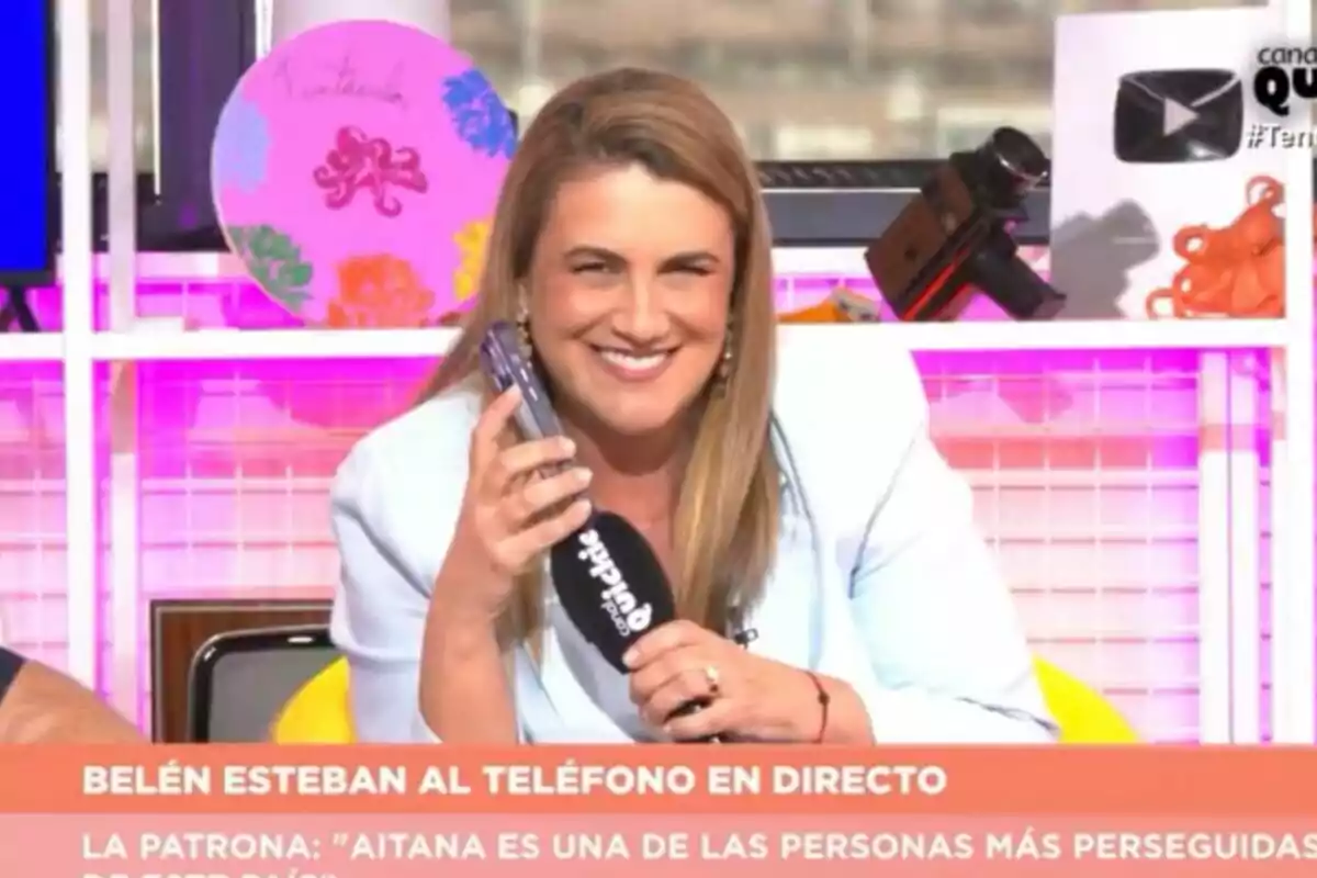 Carlota Corredera sonriente en 'Tentáculos' sosteniendo un teléfono móvil y un micrófono mientras está sentada en un set colorido con objetos decorativos al fondo