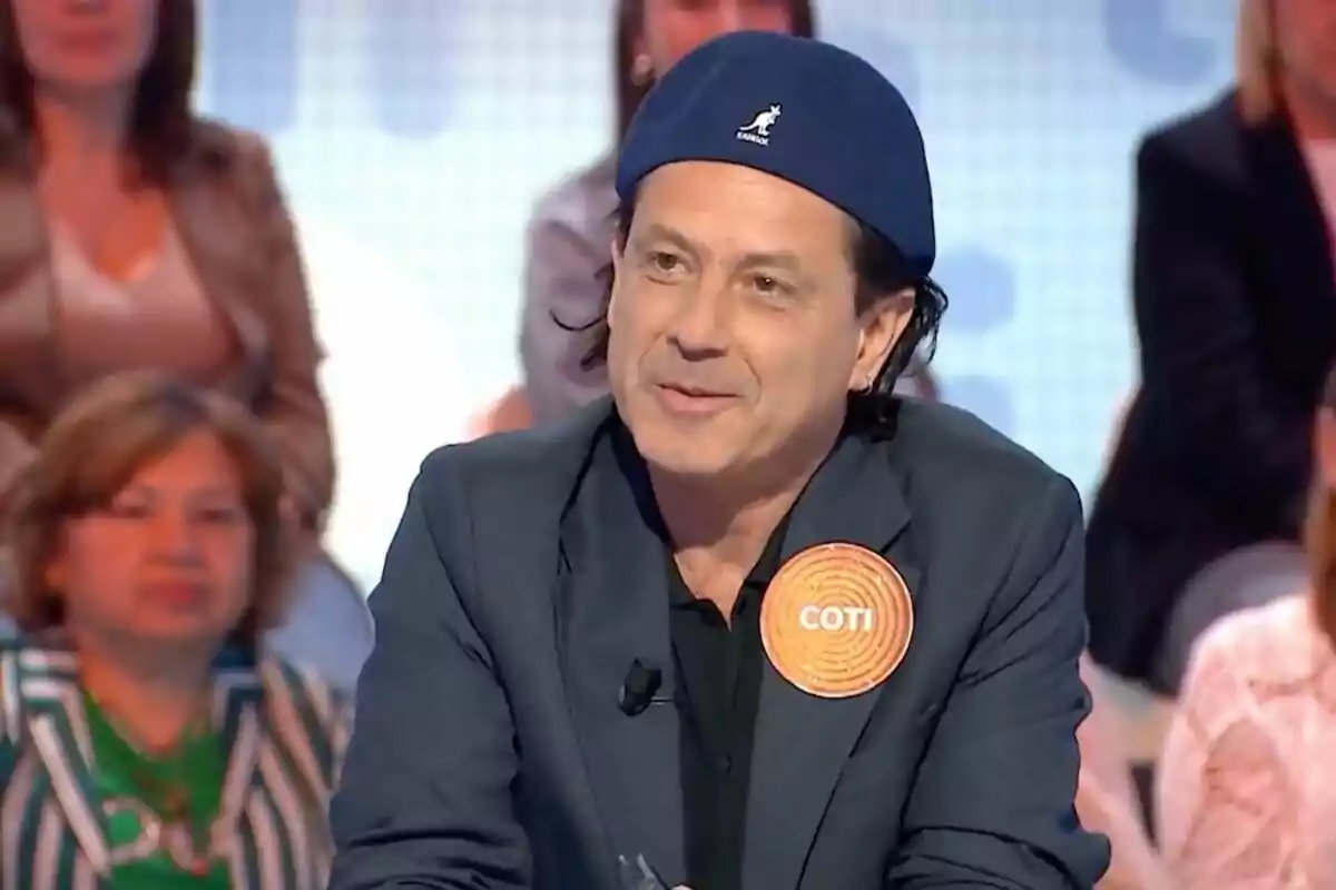 Coti con boina azul y chaqueta oscura participando en 'Pasapalabra' con público de fondo y una etiqueta que dice COTI en el pecho