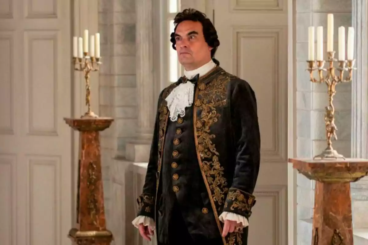 Don Hernando con peluca y traje de época elegante de pie en una habitación decorada con candelabros y columnas en escena de la serie 'Valle Salvaje'. Don Hernando con peluca y traje de época elegante de pie en una habitación decorada con candelabros y columnas en escena de la serie 'Valle Salvaje'.