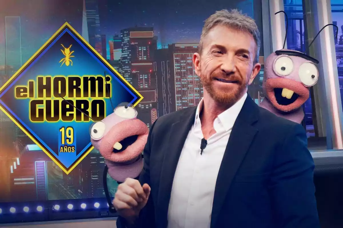 Pablo Motos sonriente con barba y traje oscuro está acompañado por dos marionetas moradas con ojos grandes y dientes salidos, al fondo se ve el logo de El Hormiguero y un fondo de ciudad iluminada