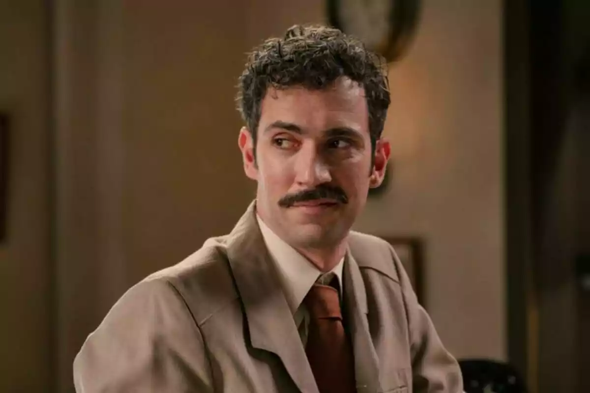 El actor Fernando Moreno con bigote con traje beige y corbata marrón sentado en una habitación iluminada mirando hacia un lado. El actor Fernando Moreno con bigote con traje beige y corbata marrón sentado en una habitación iluminada mirando hacia un lado.