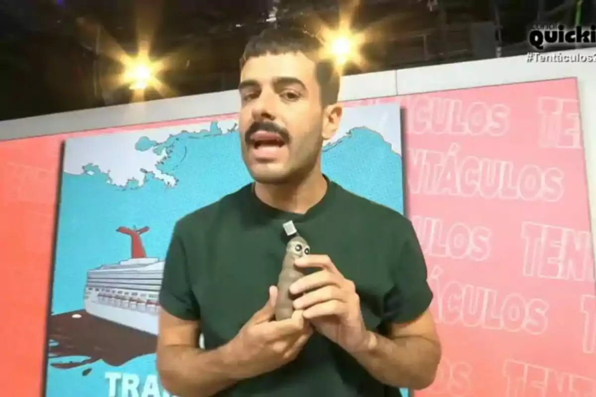 Germán González amb bigoti i samarreta verda sosté un objecte petit amb ulls mentre està dret davant d’un fons acolorit amb lletres i una il·lustració d’un creuer amb tentacles. Germán González amb bigoti i samarreta verda sosté un objecte petit amb ulls mentre està dret davant d’un fons acolorit amb lletres i una il·lustració d’un creuer amb tentacles.