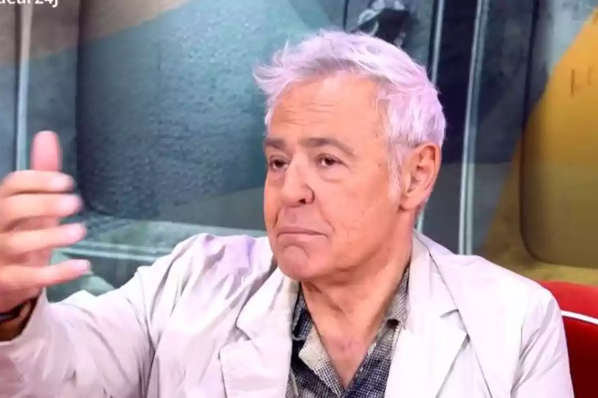 Home de cabells canosos i expressió seriosa assegut en un estudi de televisió amb el fons desenfocat i vestint una jaqueta clara