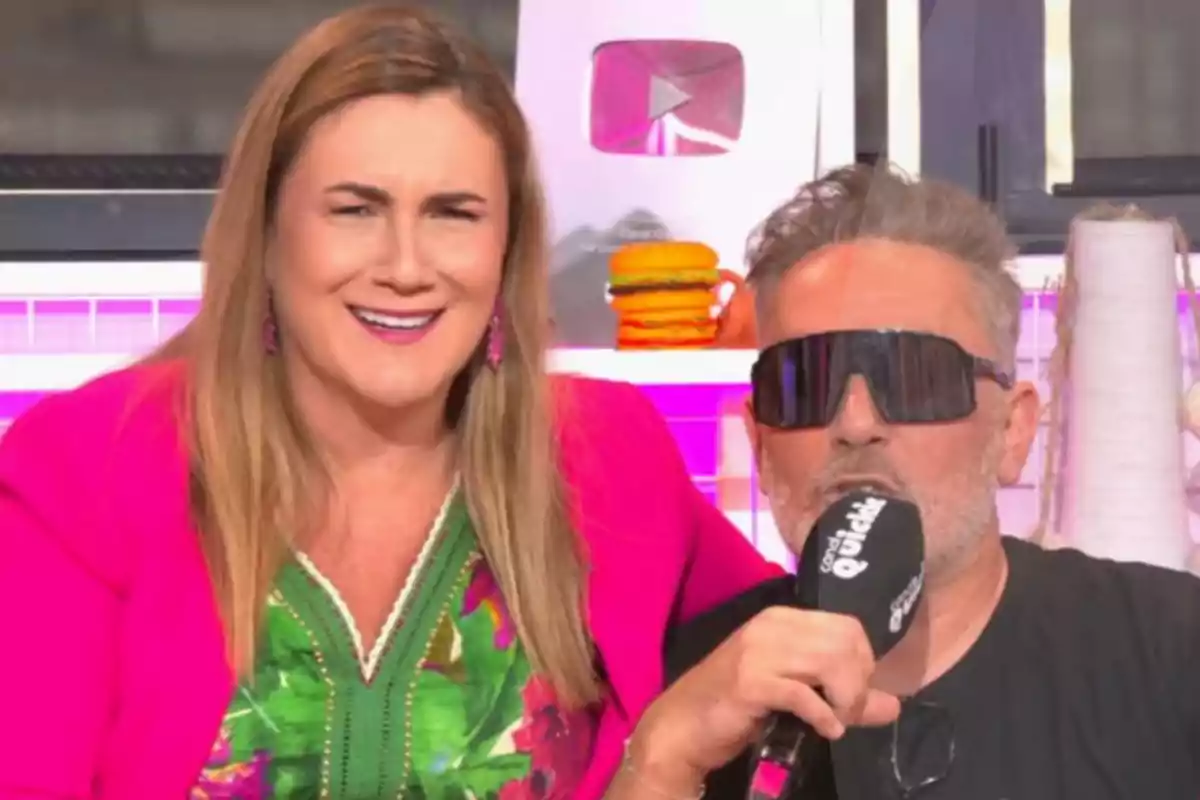 Carlota Corredera y Kiko Hernández en un set de grabación, una mujer sonriente con chaqueta rosa y un hombre con gafas oscuras sosteniendo un micrófono, al fondo un botón de YouTube plateado y decoración colorida. Carlota Corredera y Kiko Hernández en un set de grabación, una mujer sonriente con chaqueta rosa y un hombre con gafas oscuras sosteniendo un micrófono, al fondo un botón de YouTube plateado y decoración colorida.