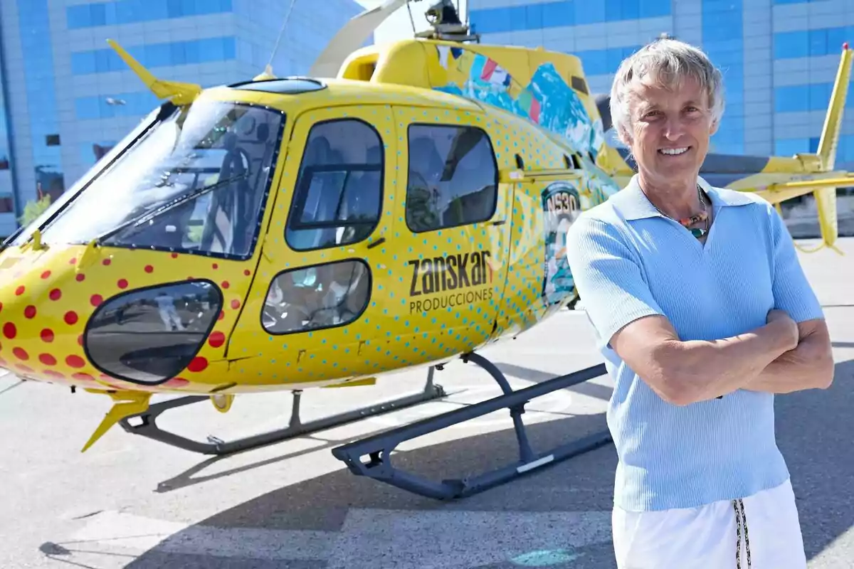 Jesús Calleja de cabello canoso y camiseta azul claro posando sonriente frente a un helicóptero amarillo decorado con puntos de colores y el logo de una productora, con un edificio moderno de fondo ante el estreno de Volando voy, volando vengo.