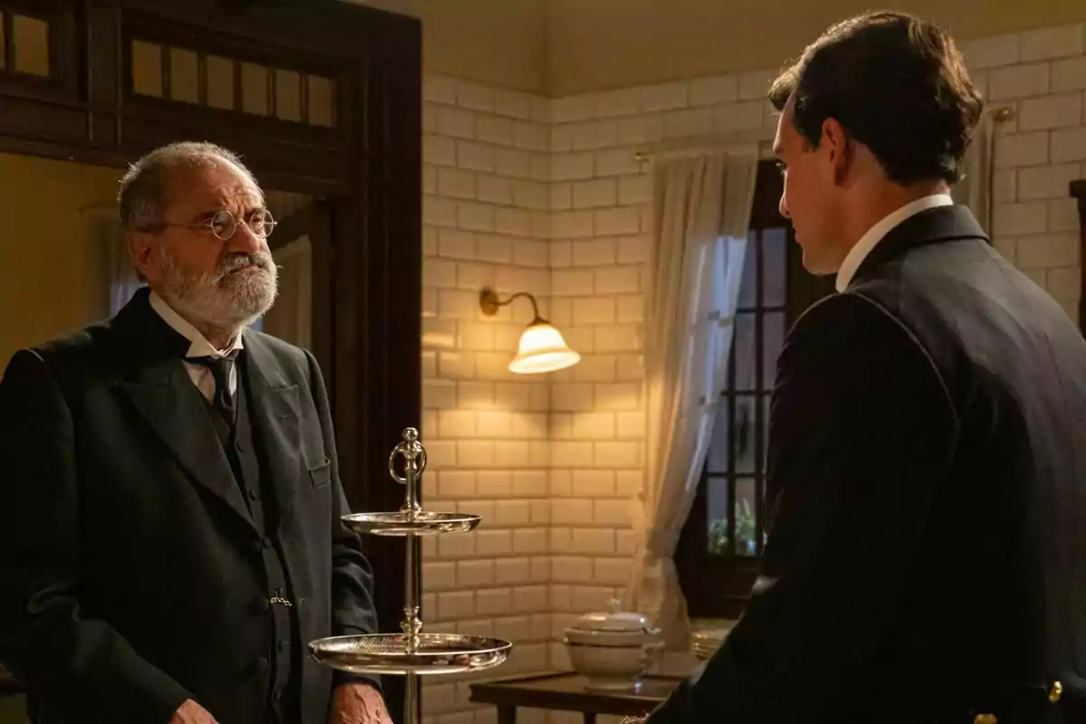 Joaquín Climent i Xavi Lock, elegantment vestits, conversen en una habitació amb parets de rajoles blanques i llum càlida a La Promesa; un d’ells porta ulleres i barba canosa, mentre que l’altre està d’esquena. Joaquín Climent i Xavi Lock, elegantment vestits, conversen en una habitació amb parets de rajoles blanques i llum càlida a La Promesa; un d’ells porta ulleres i barba canosa, mentre que l’altre està d’esquena.