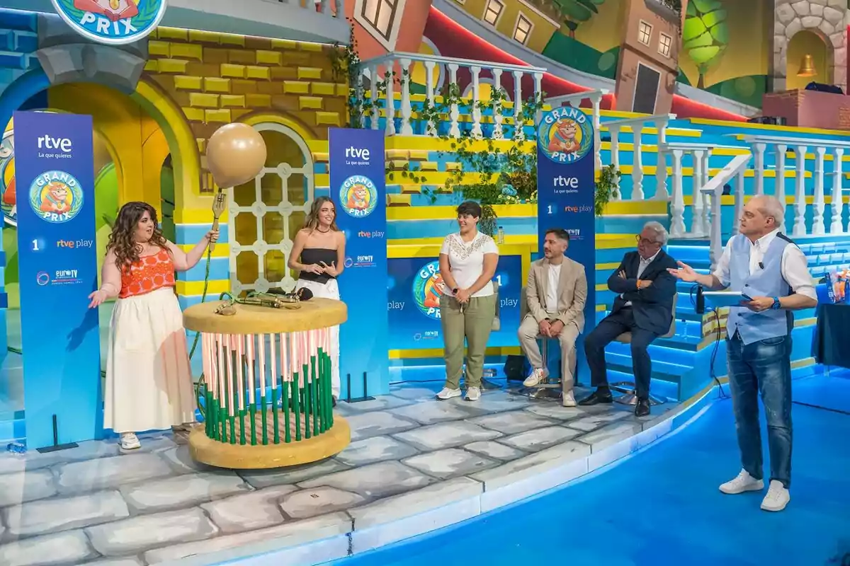 Un grup de persones participa en un programa de televisió amb una decoració colorida, la LalaChus sosté un globus gran mentre els altres observen i conversen, tots es troben en un ambient alegre i dinàmic. Un grup de persones participa en un programa de televisió amb una decoració colorida, la LalaChus sosté un globus gran mentre els altres observen i conversen, tots es troben en un ambient alegre i dinàmic.