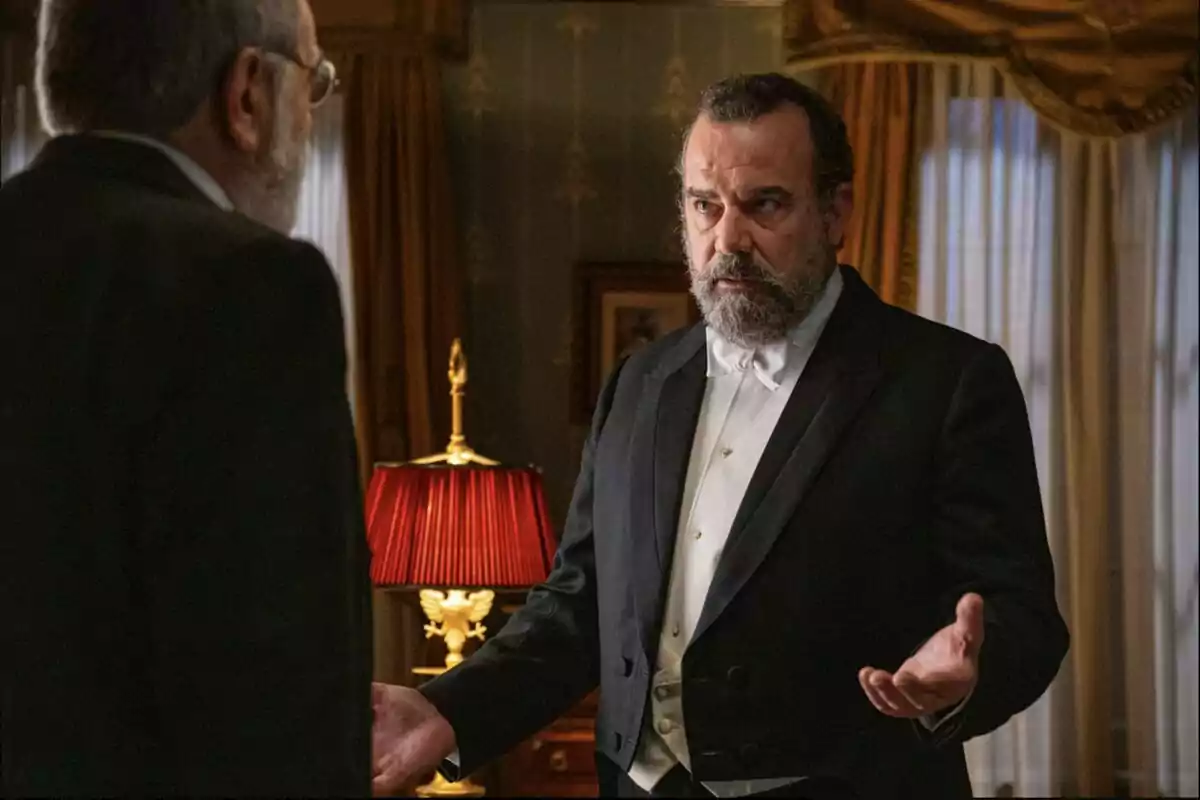 Alonso amb barba i esmòquing blanc i negre gesticulant amb les mans mentre parla amb Ròmul en una habitació elegant amb cortines daurades i una làmpada de pantalla vermella. Captura de 'La Promesa'