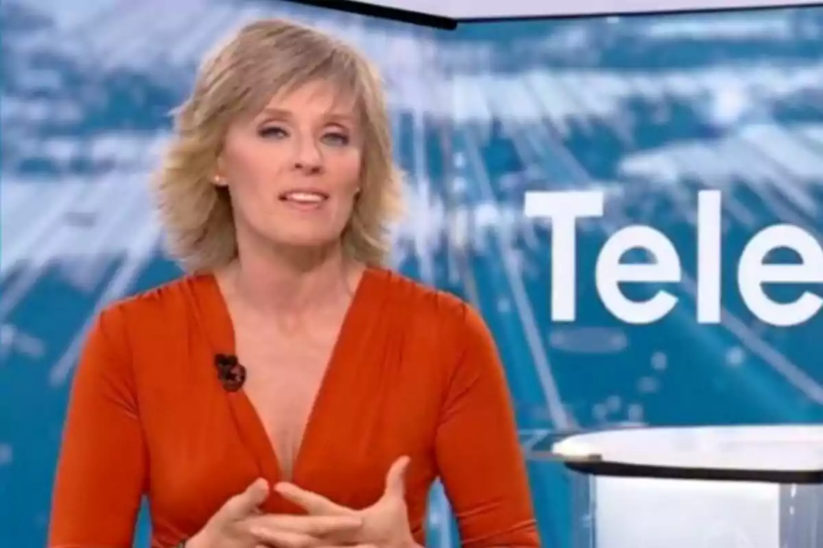Marta Carazo con vestido naranja hablando en un estudio del Telediario con fondo azul y letras blancas