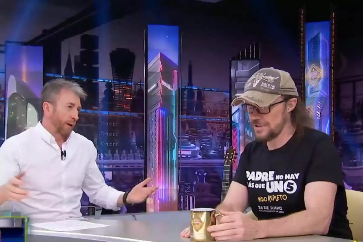 Pablo Motos i Santiago Segura conversen a 'El Hormiguero', un porta camisa blanca i l'altre samarreta negra i gorra, tots dos asseguts a una taula amb tasses daurades.