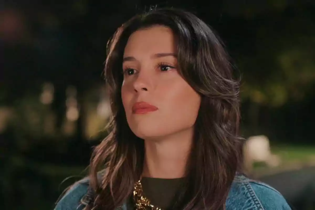 Pelin de cabello castaño largo con expresión seria, usando chaqueta de mezclilla y collar dorado, fondo desenfocado al aire libre en Una nueva vida. Pelin de cabello castaño largo con expresión seria, usando chaqueta de mezclilla y collar dorado, fondo desenfocado al aire libre en Una nueva vida.