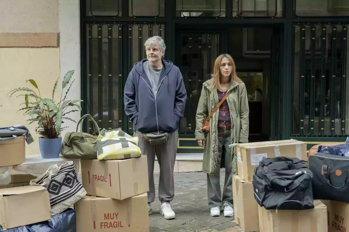 Una pareja de mediana edad, Esperanza Pedreño y Raúl Cimas, está de pie en la calle rodeada de cajas de mudanza, bolsas y una planta, frente a la entrada de un edificio en la serie 'Poquita Fe'.