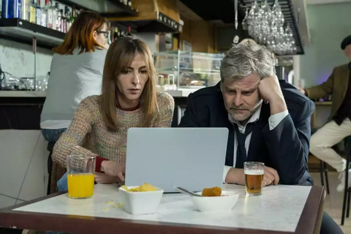 Esperanza Pedreño y Raúl Cimas sentadas en una mesa de cafetería mirando una laptop con expresión seria, hay bebidas y botanas sobre la mesa en la serie 'Poquita fe'. Esperanza Pedreño y Raúl Cimas sentadas en una mesa de cafetería mirando una laptop con expresión seria, hay bebidas y botanas sobre la mesa en la serie 'Poquita fe'.