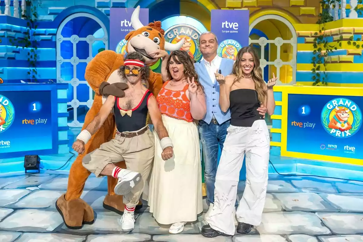 Cinc persones, Wilbur, LaLaChus, Ramón García, Ángela Fernández i una mascota disfressada de toro posen somrients en un colorit plató de televisió del programa Grand Prix, amb decoracions blaves i grogues i logotips de RTVE al fons.