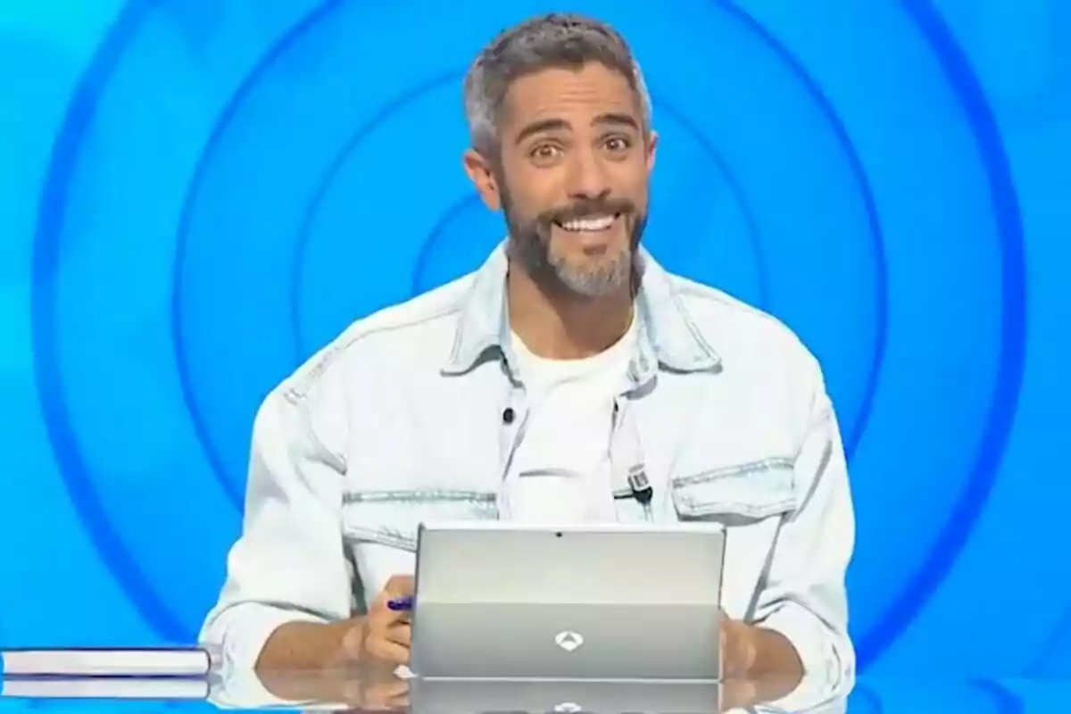 Roberto Leal sonriente con barba y chaqueta clara sentado frente a una tablet en un set de televisión con fondo azul en 'Pasapalabra'