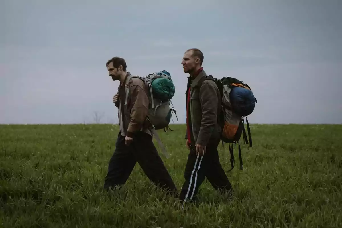 José Manuel Poga y Julián Villagrán caminan por un campo de pasto llevando mochilas grandes y equipo de campamento sobre sus espaldas bajo un cielo nublado en 33 días José Manuel Poga y Julián Villagrán caminan por un campo de pasto llevando mochilas grandes y equipo de campamento sobre sus espaldas bajo un cielo nublado en 33 días