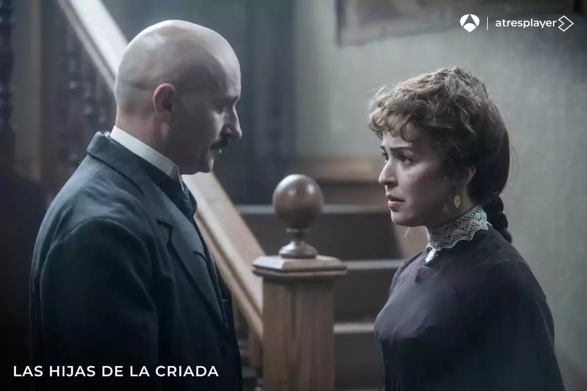 Alain Hernández calvo con bigote y Verónica Sánchez de cabello recogido con expresión seria se miran frente a una escalera de madera en una escena de época en Las hijas de la criada Alain Hernández calvo con bigote y Verónica Sánchez de cabello recogido con expresión seria se miran frente a una escalera de madera en una escena de época en Las hijas de la criada