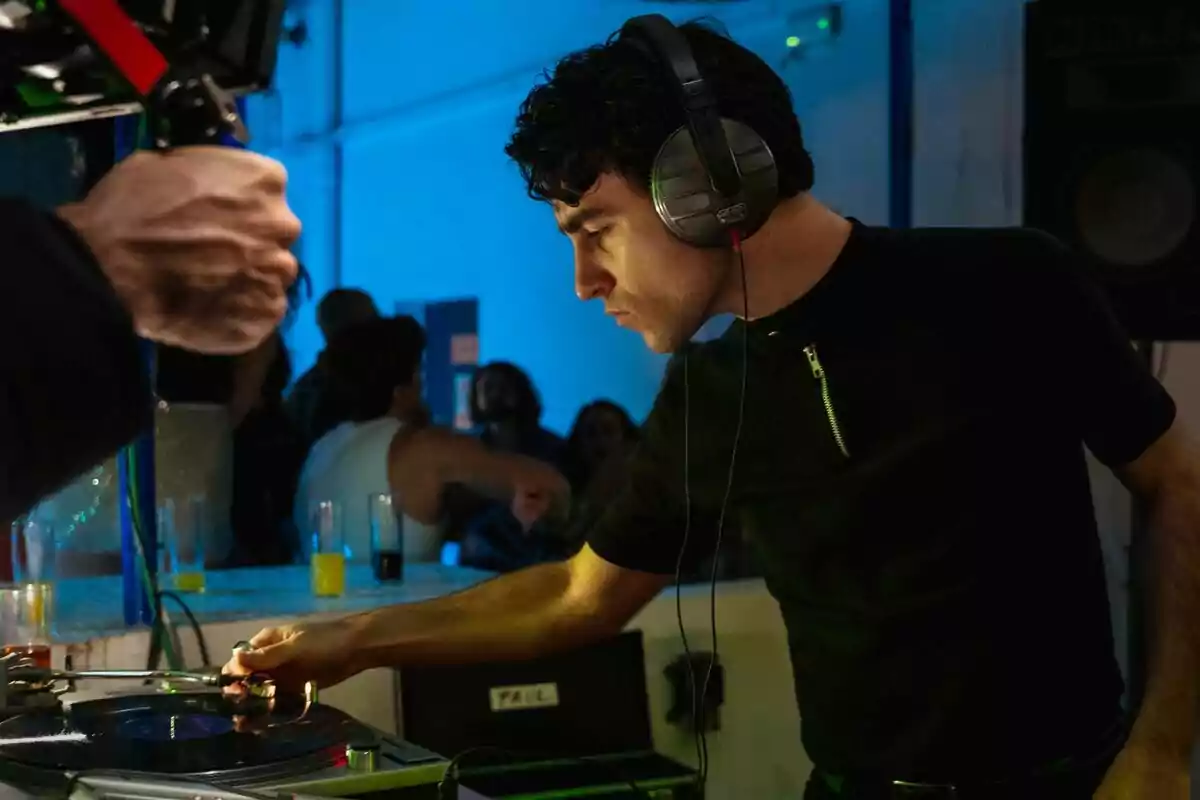 Álex Monner de DJ amb auriculars mescla música en un tocadiscs en una festa amb llums blaves i persones al fons a La Ruta 2