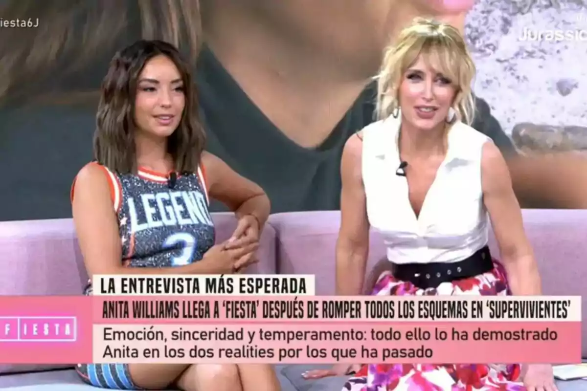 Dos mujeres sentadas en un sofá durante un programa de Fiesta, Anita Williams un vestido sin mangas con la palabra LEGEND y Emma García viste blusa blanca y falda colorida, ambas sonríen mientras aparece un texto en pantalla sobre una entrevista esperada. Dos mujeres sentadas en un sofá durante un programa de Fiesta, Anita Williams un vestido sin mangas con la palabra LEGEND y Emma García viste blusa blanca y falda colorida, ambas sonríen mientras aparece un texto en pantalla sobre una entrevista esperada.