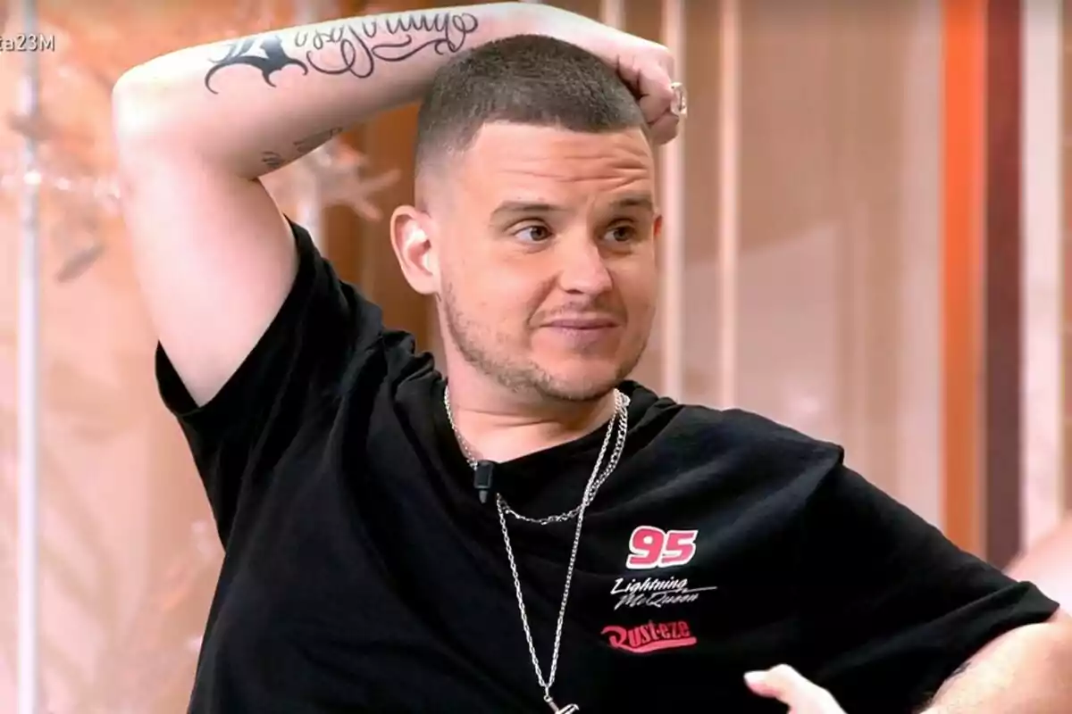 Arkano con cabello corto y barba incipiente sentado con el brazo levantado mostrando un tatuaje en el antebrazo y vistiendo una camiseta negra con el número 95 y cadenas plateadas en Fiesta. Arkano con cabello corto y barba incipiente sentado con el brazo levantado mostrando un tatuaje en el antebrazo y vistiendo una camiseta negra con el número 95 y cadenas plateadas en Fiesta.