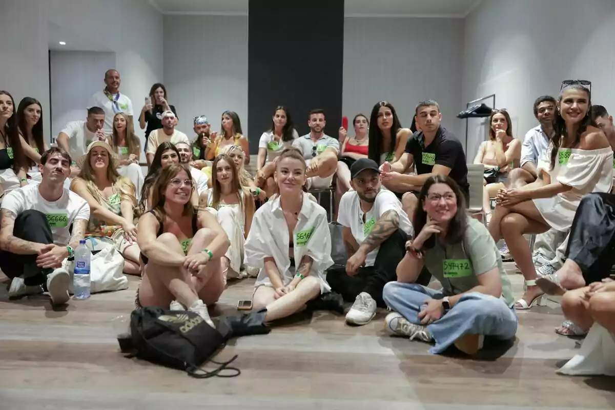 Un grupo numeroso de personas jóvenes sentadas en el suelo de una sala, algunas con camisetas blancas y etiquetas verdes, mirando hacia el frente y sonriendo en el casting de Gran Hermano Un grupo numeroso de personas jóvenes sentadas en el suelo de una sala, algunas con camisetas blancas y etiquetas verdes, mirando hacia el frente y sonriendo en el casting de Gran Hermano