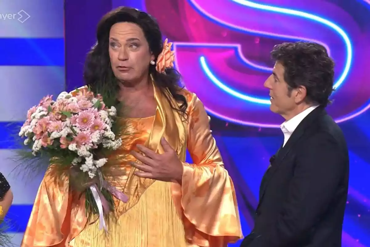 Bertín Osborne vestit amb un vestit taronja i perruca fosca sostenint un ram de flors al costat de Manel Fuentes amb vestit en un plató de Tu Cara Me Suena acolorit