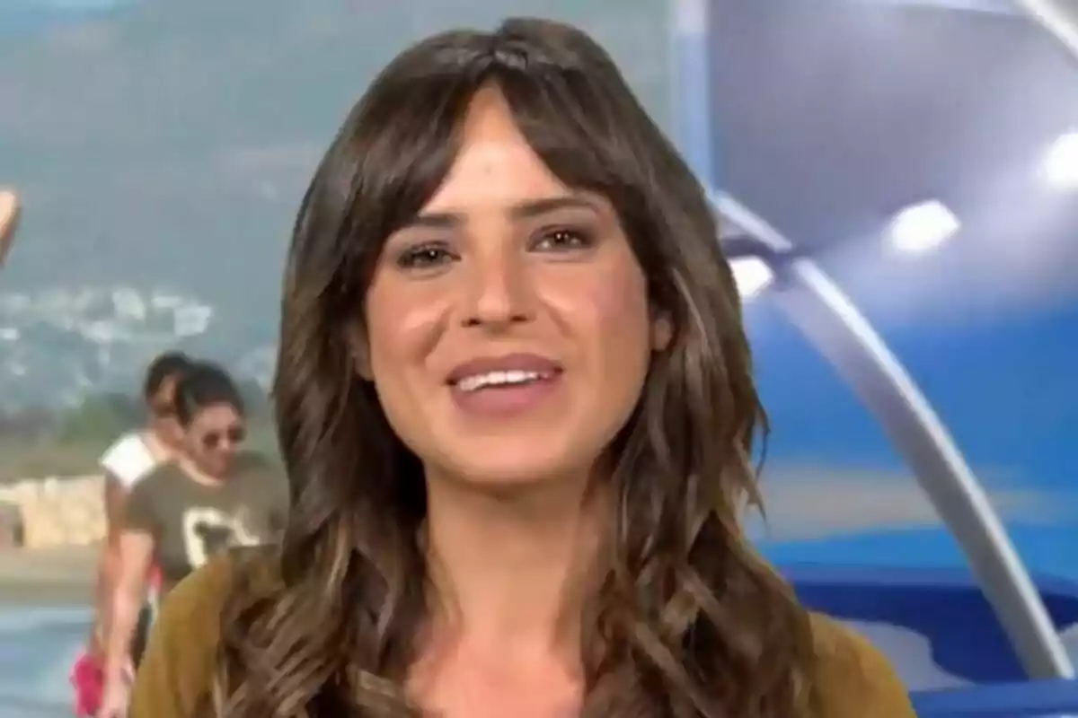 Carmen Corazzini joven de cabello castaño sonríe frente a una pantalla azul con personas desenfocadas al fondo en Informativos Telecinco
