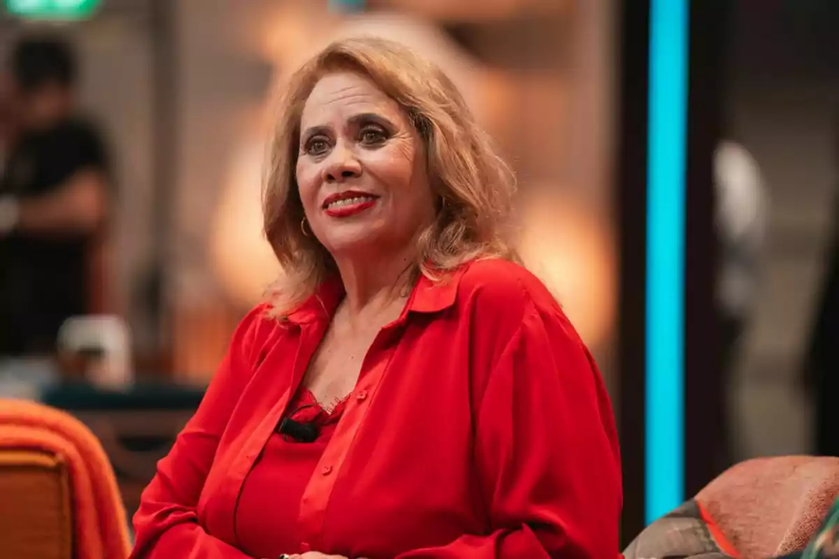 Carmina Barrios de cabello rubio y largo sentada y sonriendo, vestida con blusa roja en un ambiente interior iluminado en Pasa sin llamar Carmina Barrios de cabello rubio y largo sentada y sonriendo, vestida con blusa roja en un ambiente interior iluminado en Pasa sin llamar