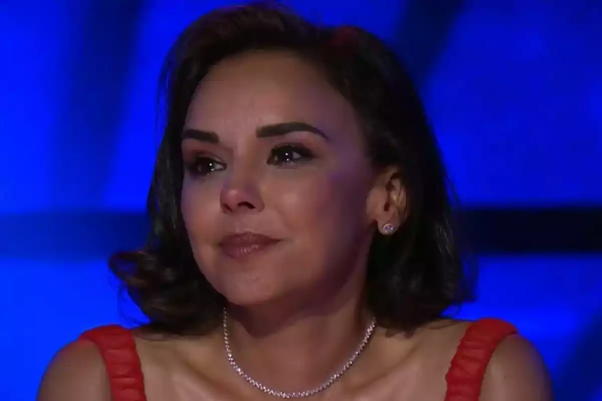 Chenoa con expresión emotiva usando un vestido rojo y collar de perlas sobre un fondo azul en Tu Cara Me Suena