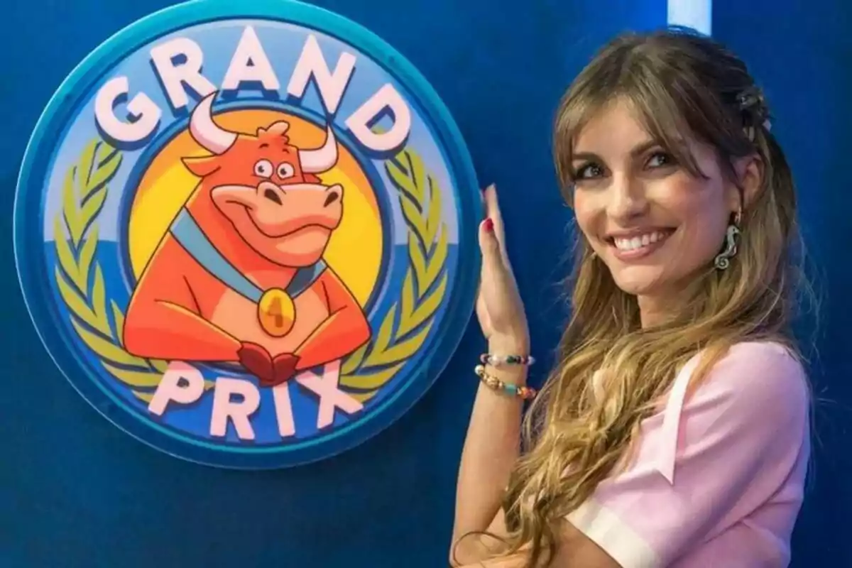Cristinini somrient amb cabell llarg i ros posa al costat d’un logotip circular del Grand Prix que mostra un toro animat amb medalla i fons blau