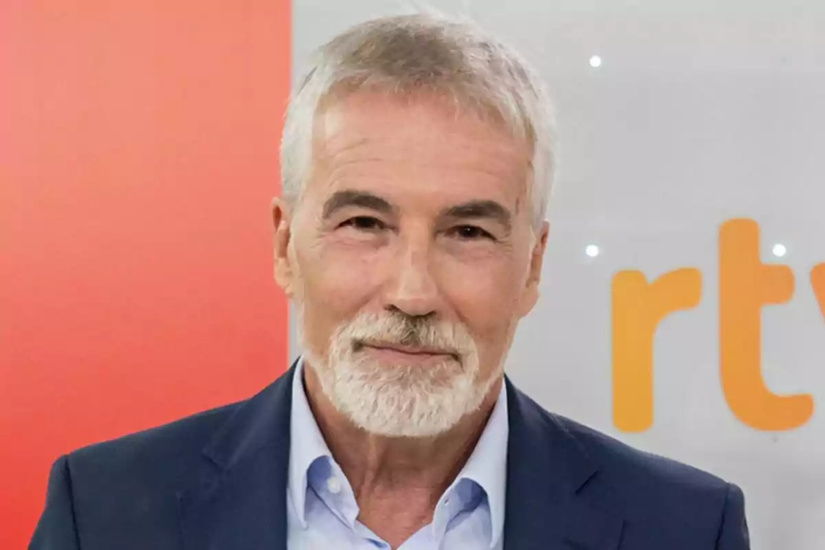 David Cantero de cabello y barba canosos con saco azul y camisa clara frente a un fondo con letras naranjas y rojo