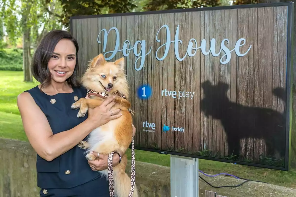 Chenoa sonriente sostiene un perro pequeño de pelaje claro frente a una pantalla que muestra el título Dog House en un entorno al aire libre Chenoa sonriente sostiene un perro pequeño de pelaje claro frente a una pantalla que muestra el título Dog House en un entorno al aire libre