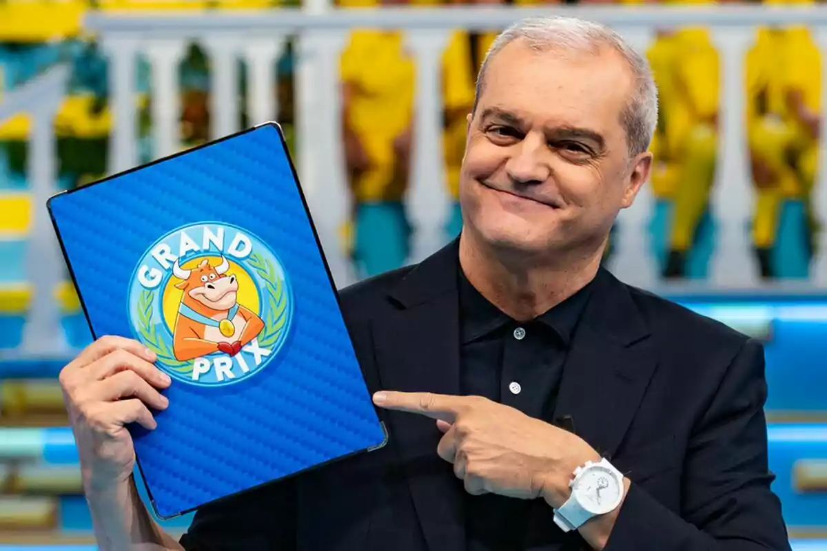 Ramón García sonriente con cabello canoso sostiene y señala una carpeta azul con el logo de Grand Prix que tiene una vaca caricaturizada