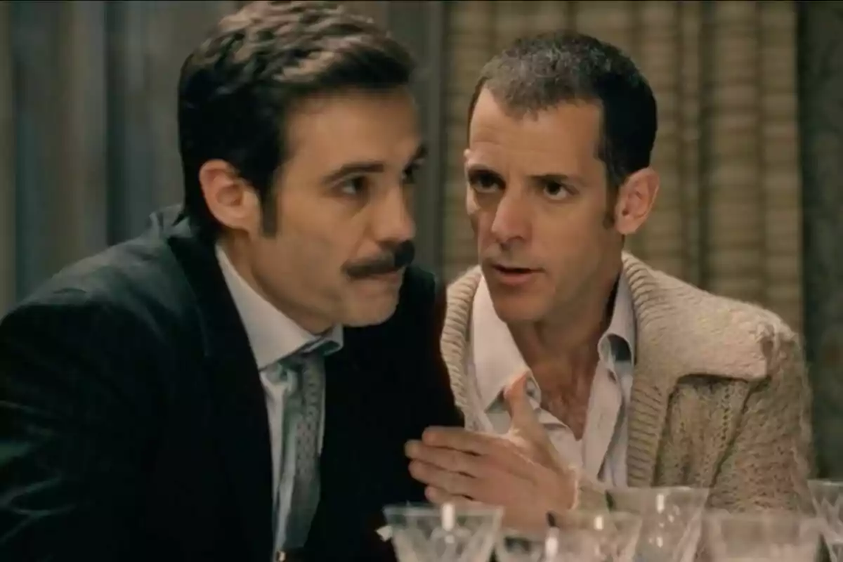 Dos hombres conversan en una mesa con copas de cristal al frente, uno de ellos lleva bigote -el cual es Guillermo Barrientos- y traje mientras el otro viste un suéter beige y parece estar hablando seriamente. Dos hombres conversan en una mesa con copas de cristal al frente, uno de ellos lleva bigote -el cual es Guillermo Barrientos- y traje mientras el otro viste un suéter beige y parece estar hablando seriamente.