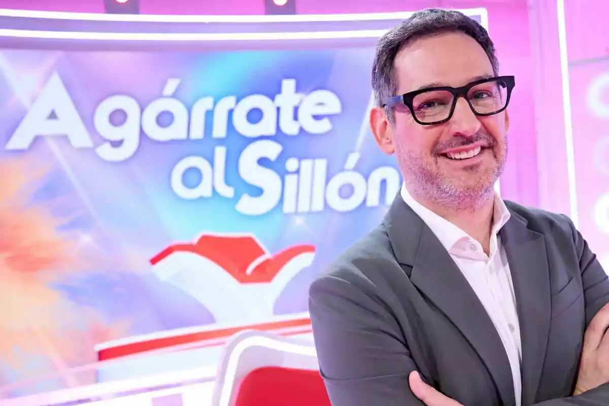 Eugeni Alemany amb ulleres i jaqueta grisa somrient davant d’un plató de televisió colorit amb el rètol Agafa’t a la Butaca al fons