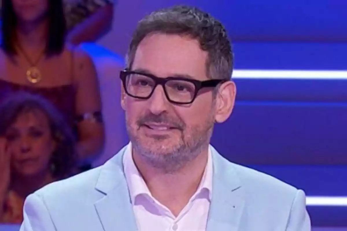 Eugeni Alemany amb ulleres i barba vestit amb jaqueta blau clar en un plató d’Agafa’t al sofà amb fons blau i públic desenfocat.