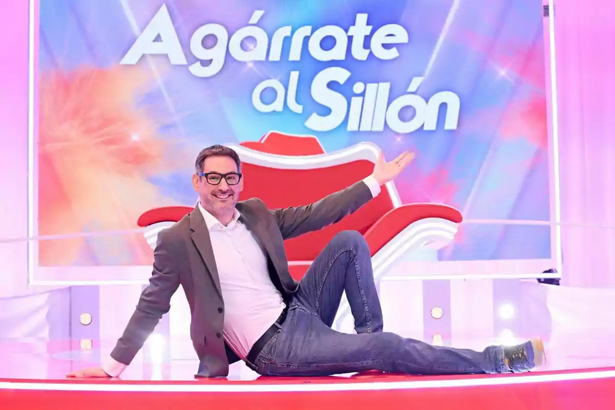 Eugeni Alemany con gafas y saco sentado en el suelo de un set de televisión colorido con un sillón rojo y un letrero de Agárrate al Sillón al fondo Eugeni Alemany con gafas y saco sentado en el suelo de un set de televisión colorido con un sillón rojo y un letrero de Agárrate al Sillón al fondo