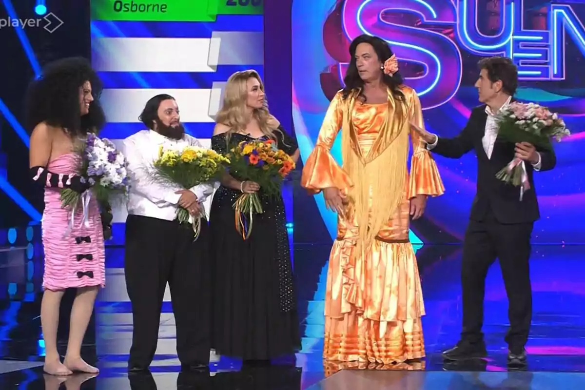 Mikel Herzog, Ana Guerra, Gisela, Bertín Osborne i Manel Fuentes en un escenari de Tu Cara Me Suena, totes sostenint rams de flors i vestides amb roba cridanera i elegant, mentre interactuen entre elles sota llums de colors. Mikel Herzog, Ana Guerra, Gisela, Bertín Osborne i Manel Fuentes en un escenari de Tu Cara Me Suena, totes sostenint rams de flors i vestides amb roba cridanera i elegant, mentre interactuen entre elles sota llums de colors.