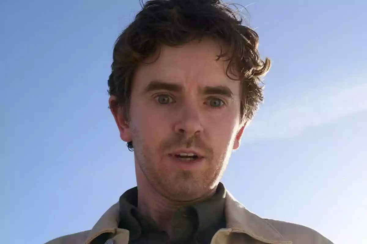 Freddie Highmore joven de cabello rizado y expresión sorprendida mirando hacia abajo con el cielo azul de fondo en Asesina a sueldo