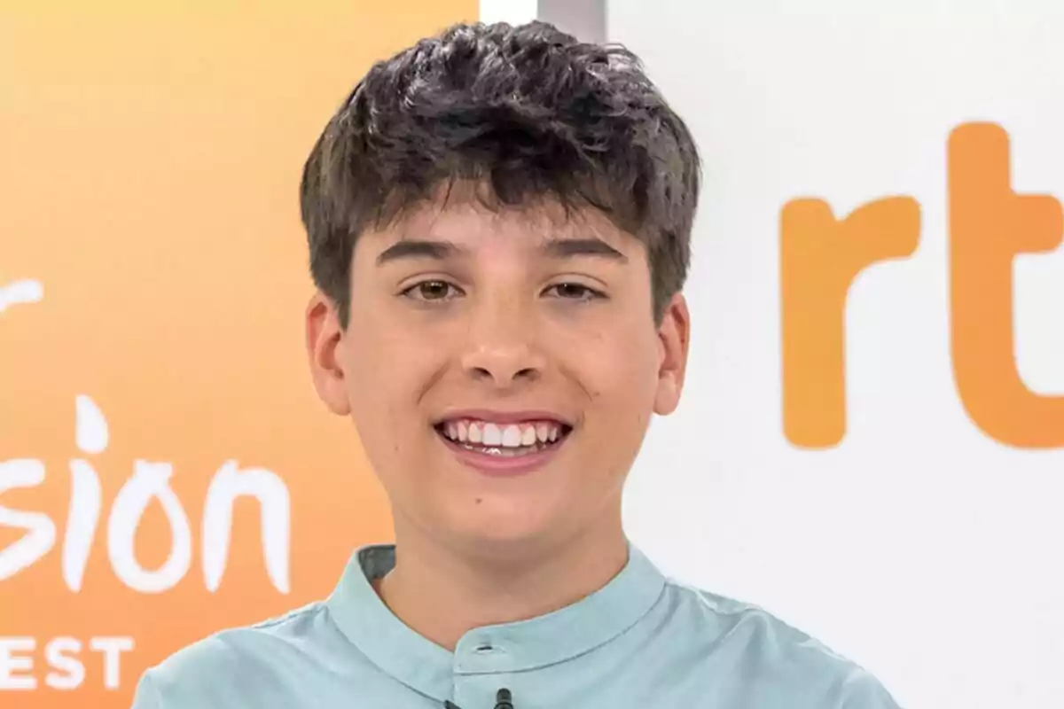 Gonzalo Pinilla somrient amb camisa celeste davant d’un fons amb logotips en tons taronja i blanc com a representant d’Eurovisió Júnior