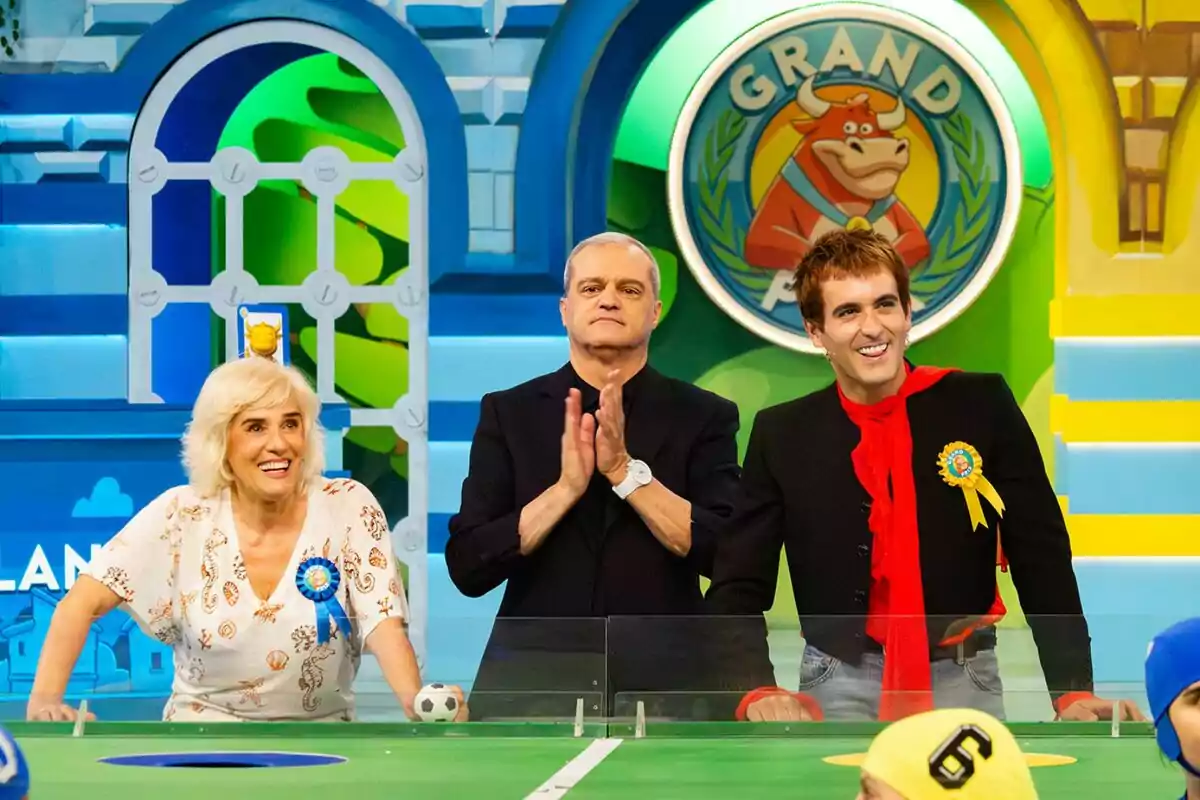 Anabel Alonso, Ramón García y Palomo Spain sonríen y posan frente a una mesa de juegos en un colorido set de El Grand Prix con un logo de una vaca al fondo Anabel Alonso, Ramón García y Palomo Spain sonríen y posan frente a una mesa de juegos en un colorido set de El Grand Prix con un logo de una vaca al fondo