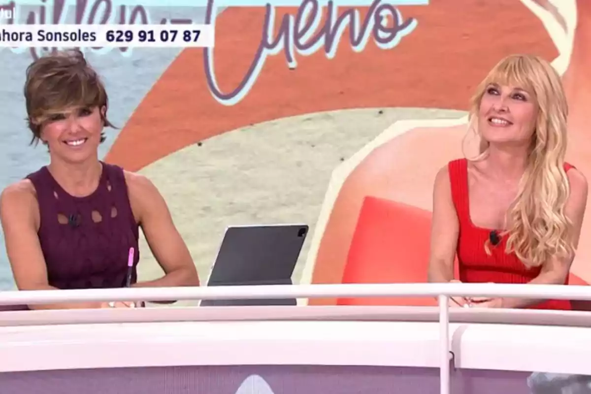 Dues dones somrients assegudes en un plató de televisió amb un fons acolorit i una tauleta sobre la taula Dues dones somrients assegudes en un plató de televisió amb un fons acolorit i una tauleta sobre la taula