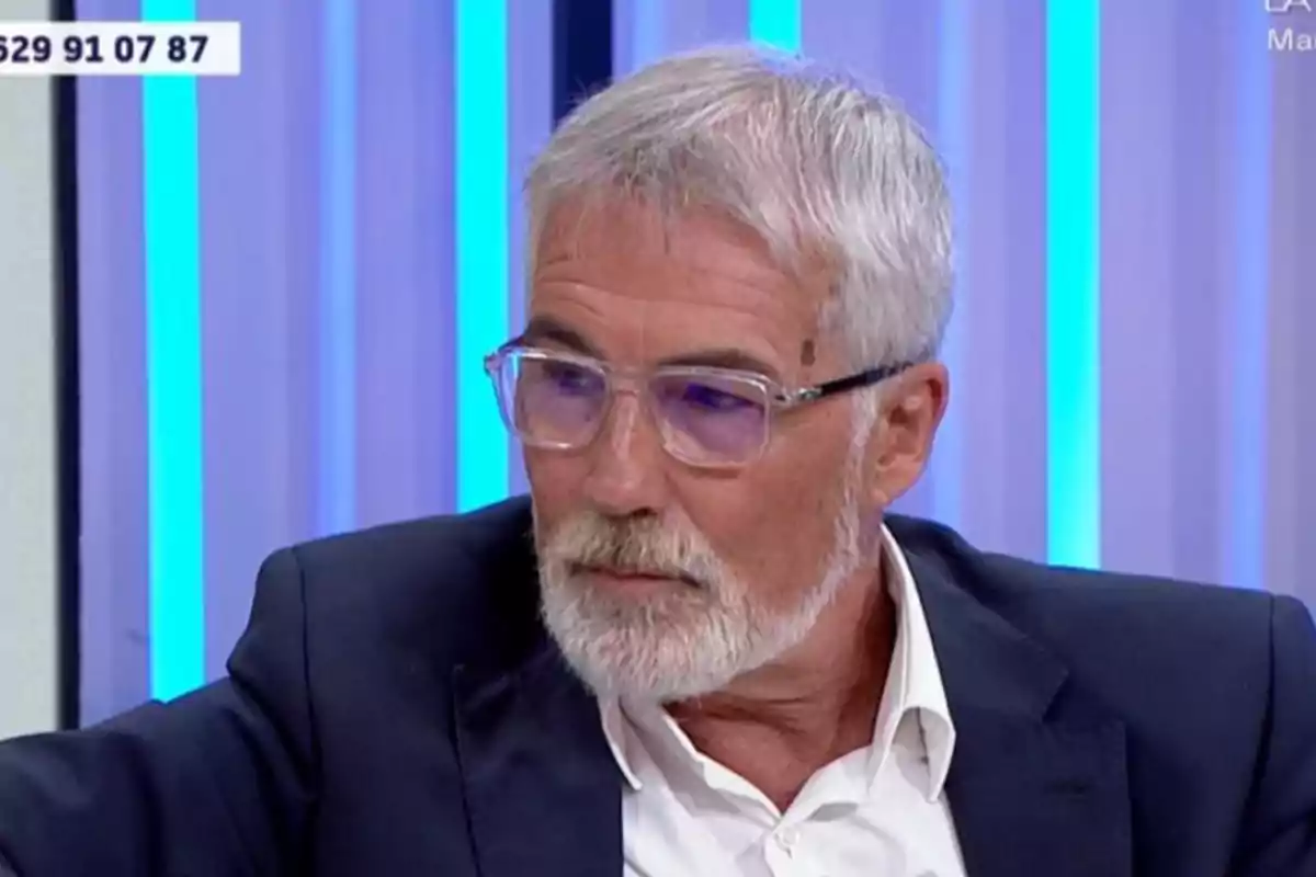 David Cantero de cabello y barba canosos con gafas transparentes y traje oscuro sentado en un estudio de televisión con luces azules de fondo
