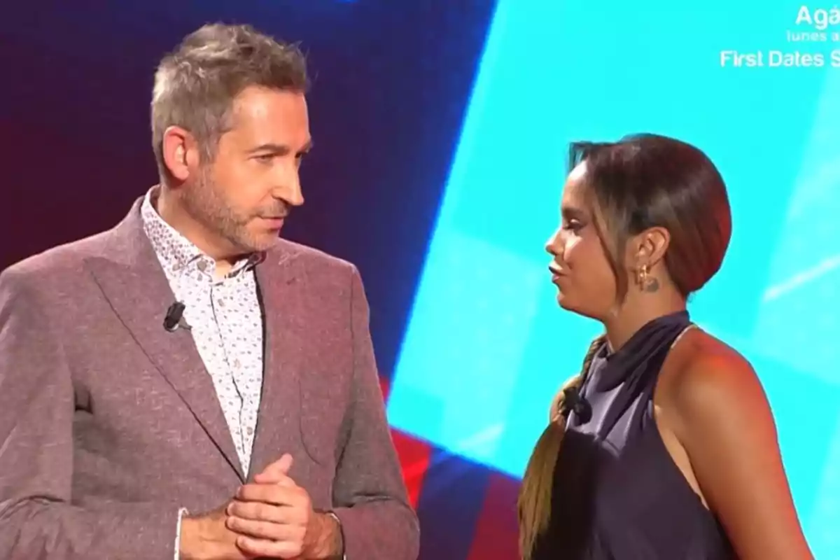 Frank Blanco con saco claro y Gloria Camila con vestido oscuro conversan en un set de televisión con fondo azul.