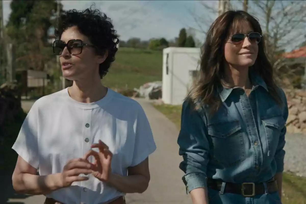 Isabel Jiménez y Blanca Romerto caminan al aire libre en un día soleado, ambas llevan gafas de sol y una de ellas viste una camisa blanca mientras la otra lleva una camisa de mezclilla en Mis Raíces Isabel Jiménez y Blanca Romerto caminan al aire libre en un día soleado, ambas llevan gafas de sol y una de ellas viste una camisa blanca mientras la otra lleva una camisa de mezclilla en Mis Raíces