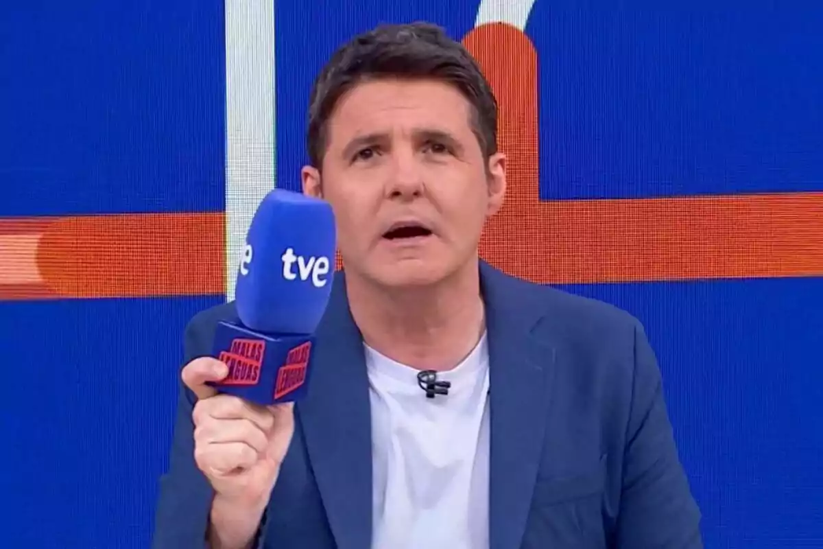 Jesús CIntora con chaqueta azul y camiseta blanca sostiene un micrófono azul con el logo de tve y la frase Malas Lenguas en un set de televisión con fondo azul y naranja