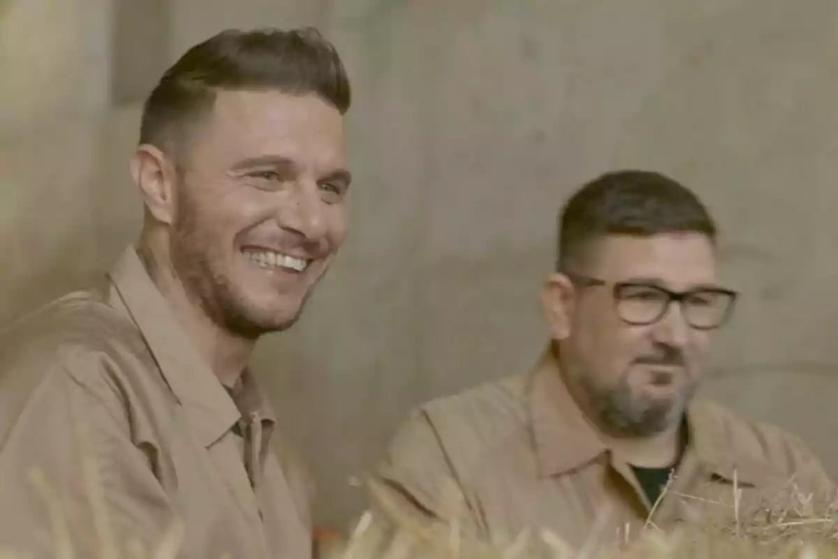 Joaquín Sánchez i Dani García amb camises beix estan asseguts junts, un d’ells somriu àmpliament i l’altre porta ulleres i barba, tots dos es troben en un entorn amb palla i fons neutre.
