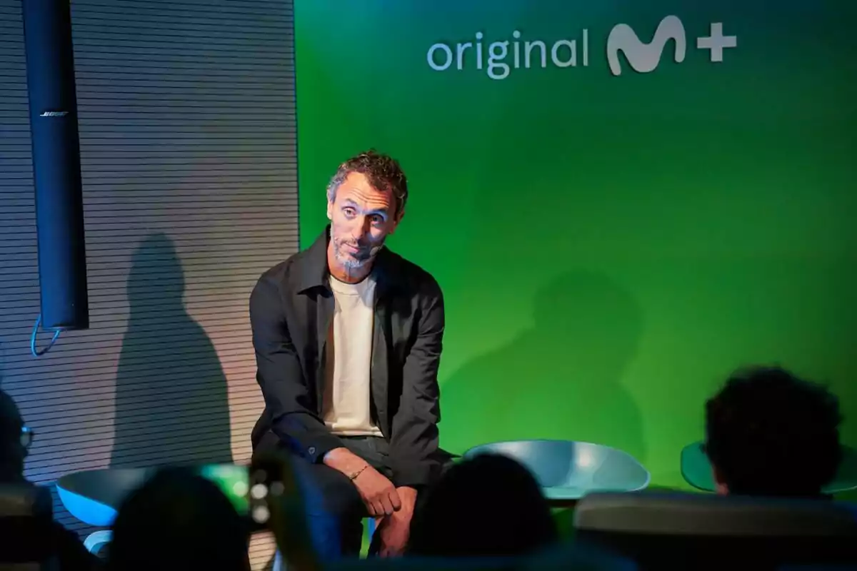Jorge Pezzi sentado frente a una pared verde con el logo de Movistar Plus y varias personas observándolo Jorge Pezzi sentado frente a una pared verde con el logo de Movistar Plus y varias personas observándolo